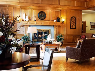 Wood-paneled communal living room with a lit fireplace, upholstered chairs, tables, lamps, and floral arrangements.