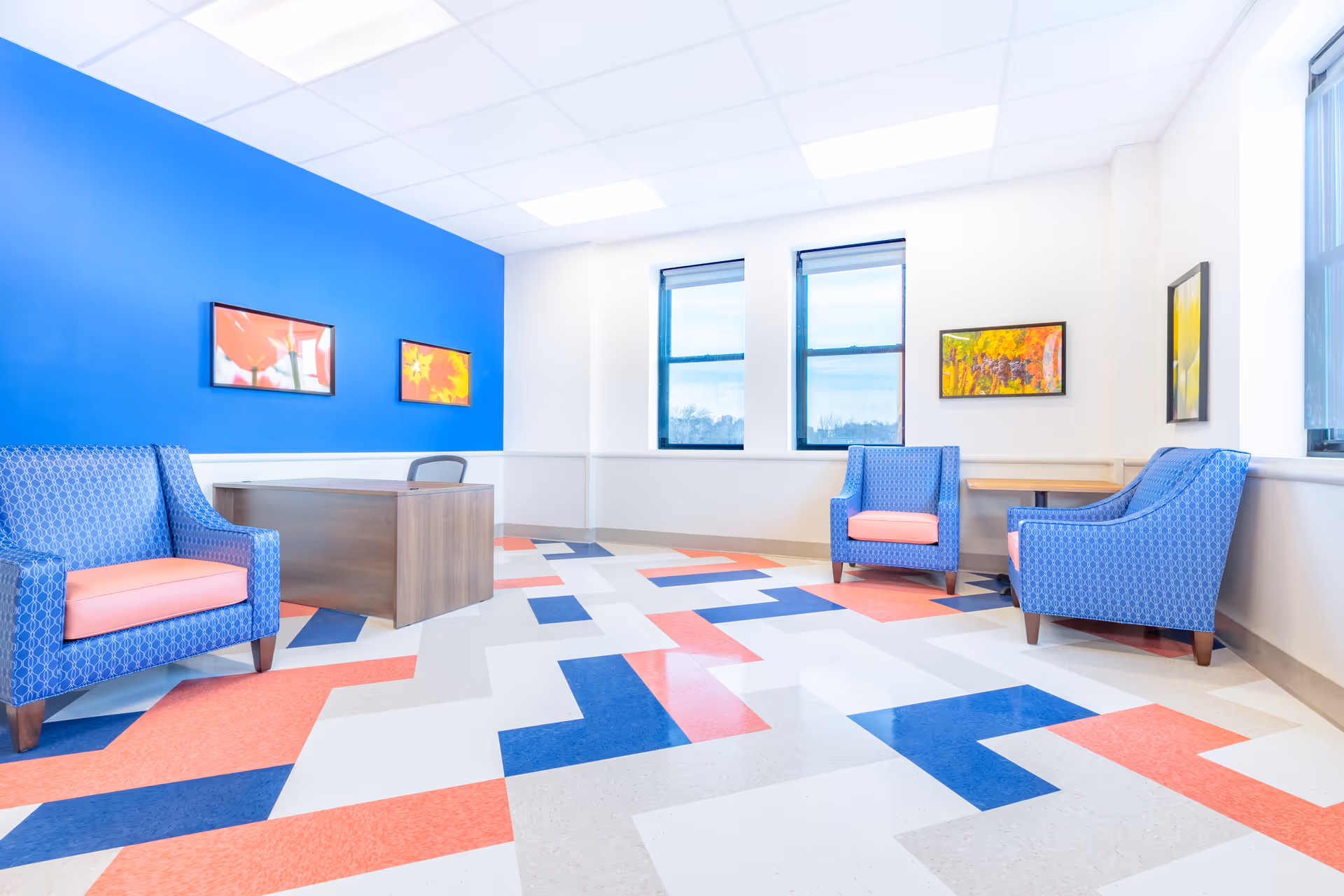 A bright and modern interior room with a blue accent wall, three blue armchairs with pink cushions, a wooden desk with a chair, and colorful abstract paintings on the walls. The floor has a geometric pattern in blue, red, and white tiles. Two large windows let in natural light.