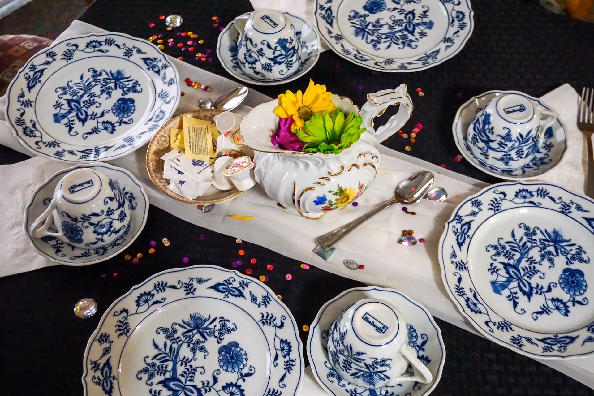 A table set with blue and white floral patterned plates and teacups, a white table runner, a small ornate white creamer filled with colorful flowers, a small dish with creamers and sugar packets, and scattered decorative sequins.