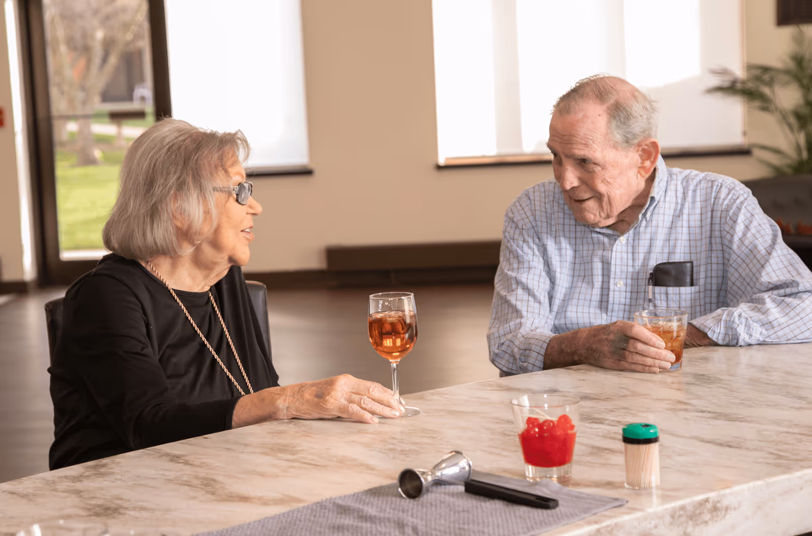 An elderly woman and an elderly man sitting at a marble countertop, engaged in conversation while holding drinks. The woman has gray hair and is wearing glasses and a black top, while the man is wearing a checkered shirt. On the countertop, there is a glass with red cherries, a jigger, a muddler, and a container of toothpicks. The background shows large windows with natural light coming in.