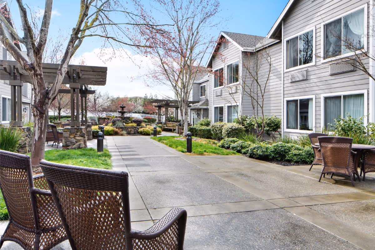 Paved courtyard at a senior living facility with wicker chairs and tables, pergolas, landscaped beds and a central fountain beside a two-story gray building.