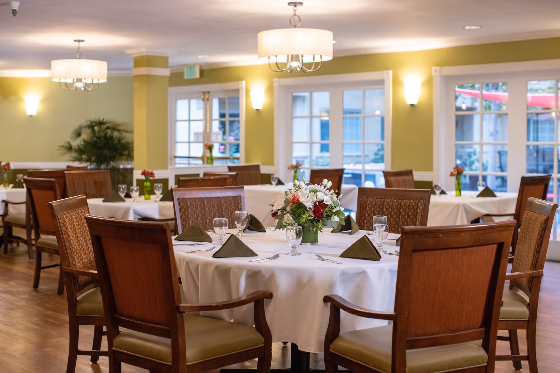 A dining room in a senior living facility with round tables covered in white tablecloths, each set with folded green napkins, glassware, and silverware. The room has wooden chairs with cushioned seats, green walls, large windows letting in natural light, and ceiling light fixtures. A floral centerpiece is on the closest table.