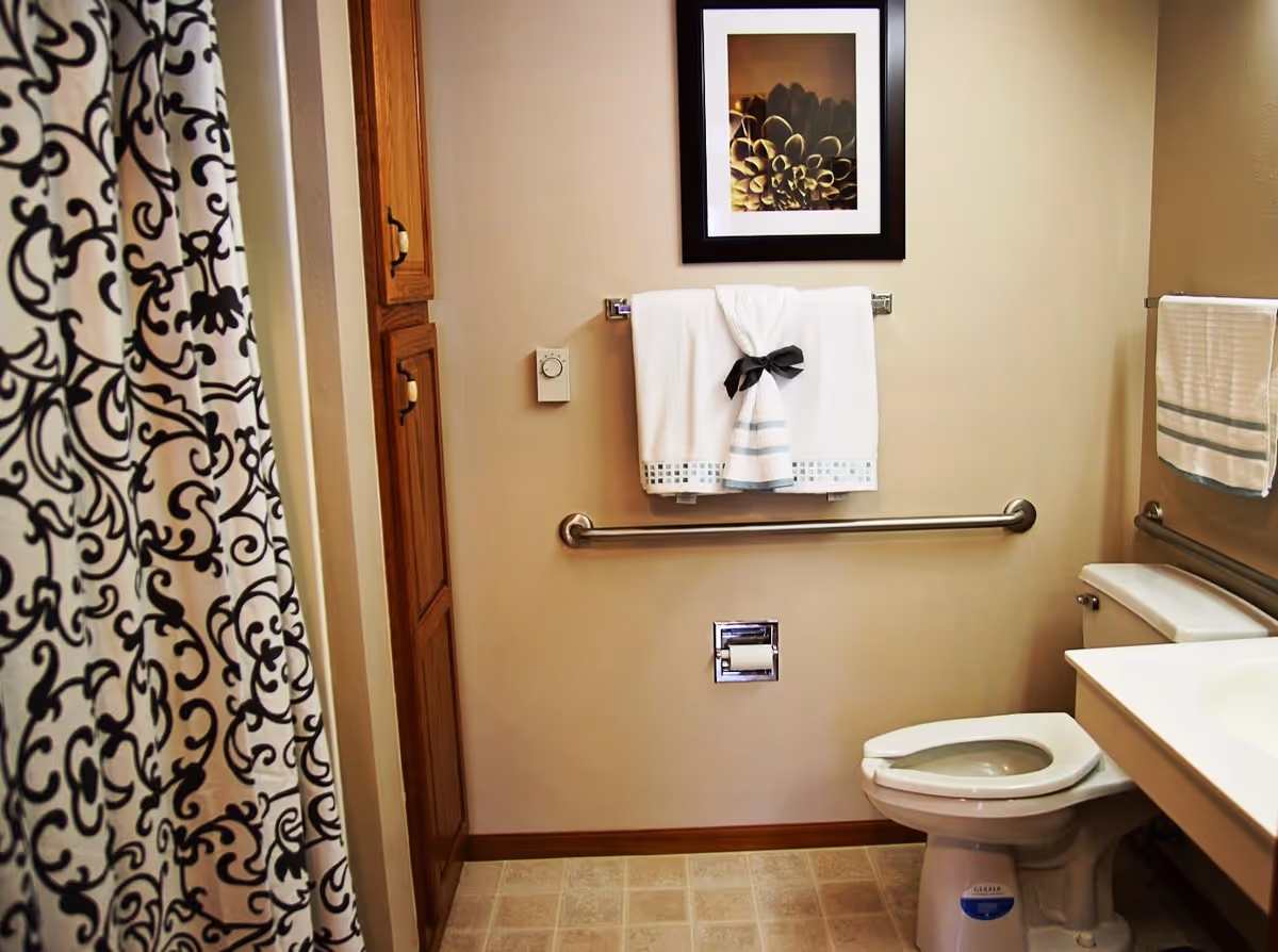 A bathroom with a toilet, a sink with a countertop, a towel rack with white towels, a decorative framed picture on the wall, a patterned shower curtain, and wooden cabinets.