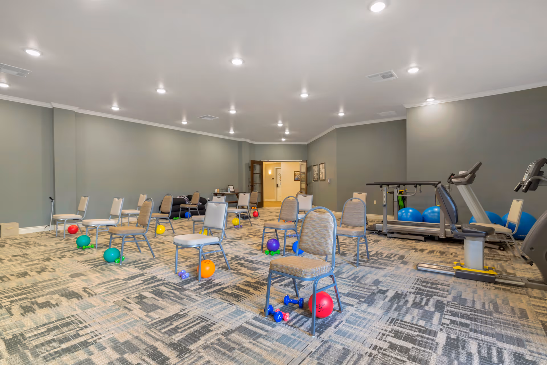 A spacious exercise room with several chairs spaced apart, each accompanied by colorful exercise balls and small dumbbells on a patterned carpet. The room has gray walls, recessed ceiling lights, and exercise equipment including treadmills and large blue exercise balls along one wall.
