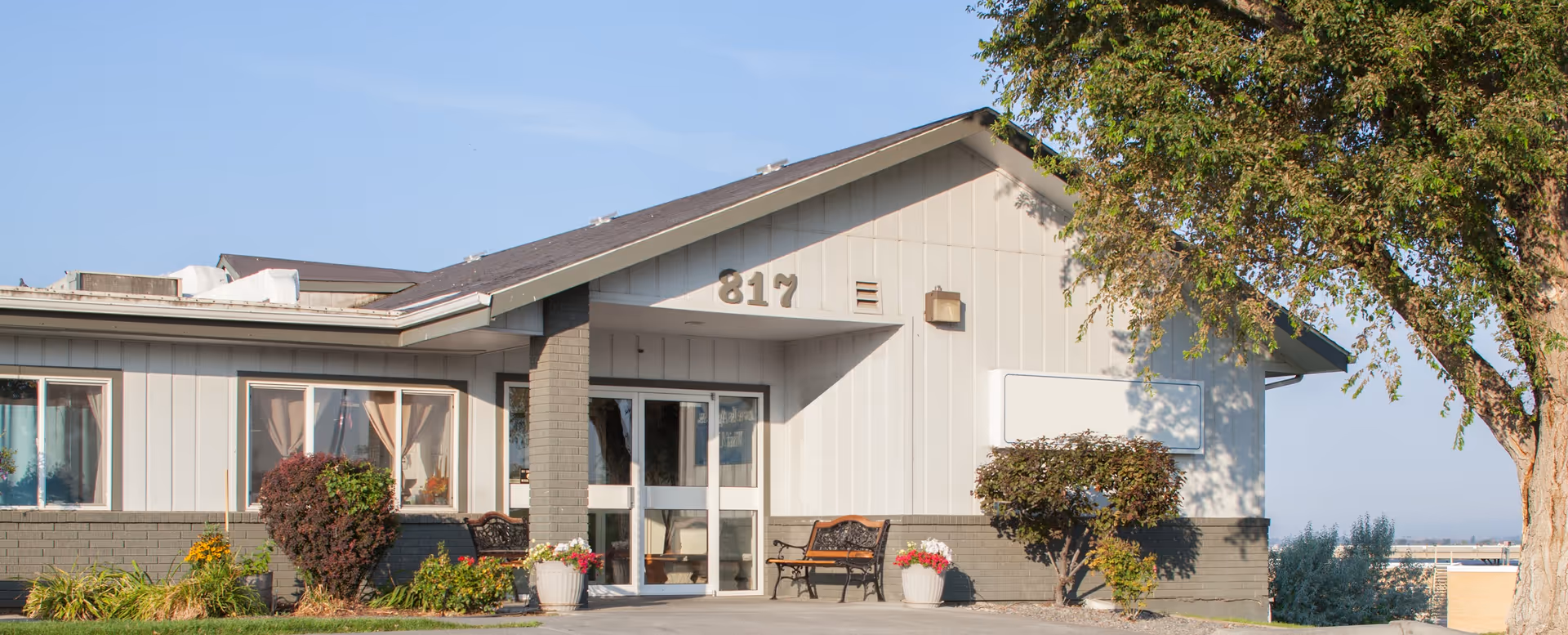 Exterior view of a single-story building with the number 817 displayed near the roof peak. The building has large windows with curtains, a glass entrance door, two benches, potted plants, and some bushes. A tree is visible on the right side, and the sky is clear and blue.