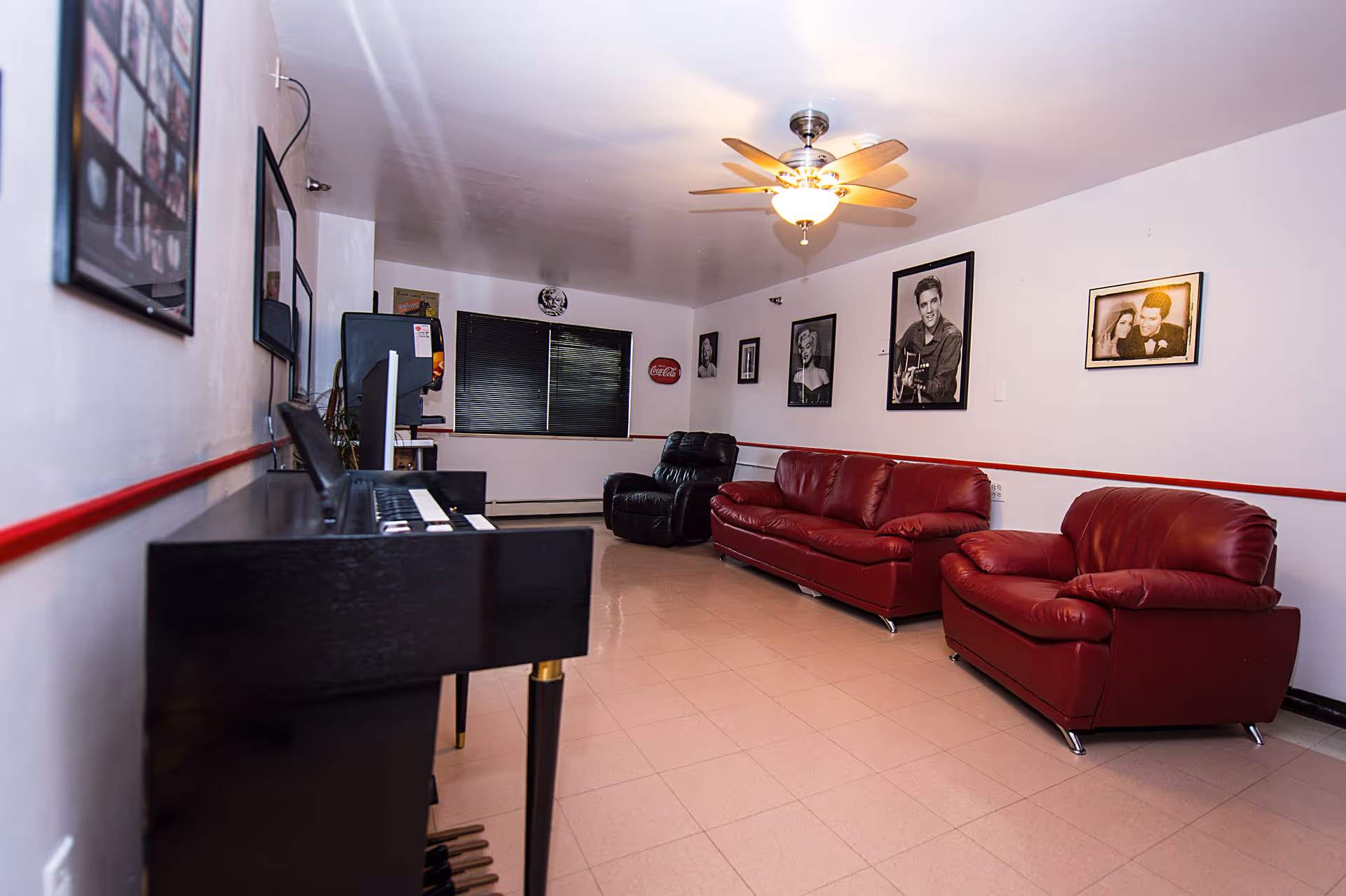 Long lounge room with red leather sofas, a ceiling fan, a piano, and framed black-and-white portraits on the walls.