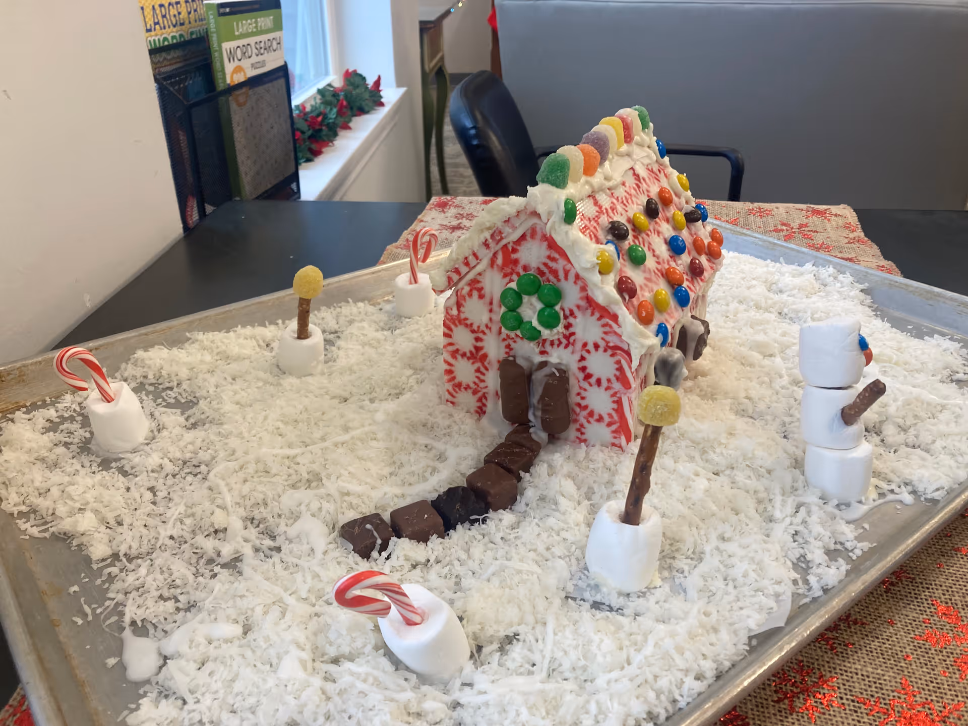 A festive candy-covered gingerbread house on a tray surrounded by shredded coconut 'snow' and marshmallow and candy decorations on a table.
