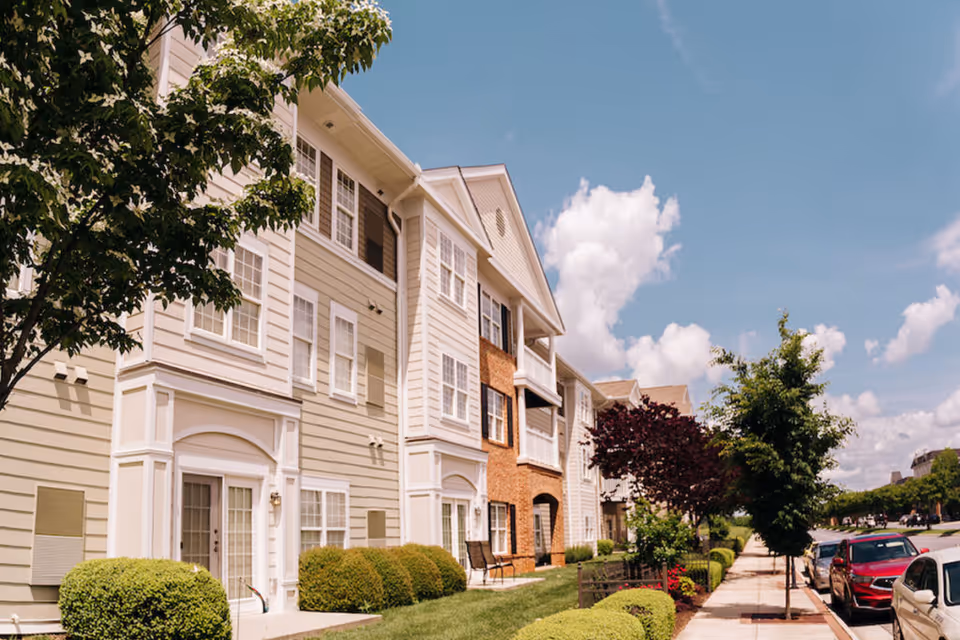 Exterior view of a multi-story residential building with beige and brick facades, surrounded by neatly trimmed bushes and trees along a sidewalk with parked cars under a partly cloudy sky.