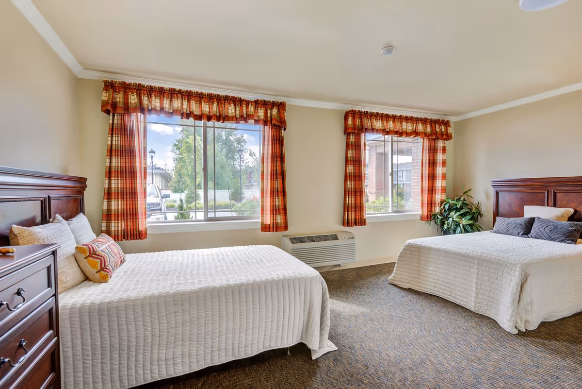 A bright bedroom with two beds featuring wooden headboards and white bedspreads. Each bed has decorative pillows. There are two large windows with red and orange plaid curtains letting in natural light. A wooden dresser is visible on the left side, and a green potted plant is placed near the right window. The room has beige walls and a carpeted floor.