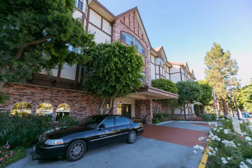 Exterior view of a multi-story assisted living facility with Tudor-style architecture, featuring brick and stucco walls, large windows, and a covered entrance. A black sedan is parked in front of the entrance, surrounded by green trees and flowering bushes under a clear blue sky.
