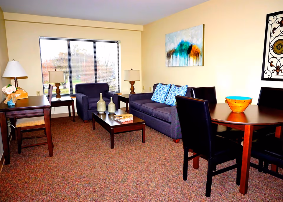 A cozy living room with a large window showing an outdoor view. The room features a dark blue armchair and sofa with blue patterned cushions, a wooden coffee table with decorative vases and books, two side tables with lamps, a wooden dining table with four black chairs, and wall art including a colorful abstract painting and a decorative metal piece.