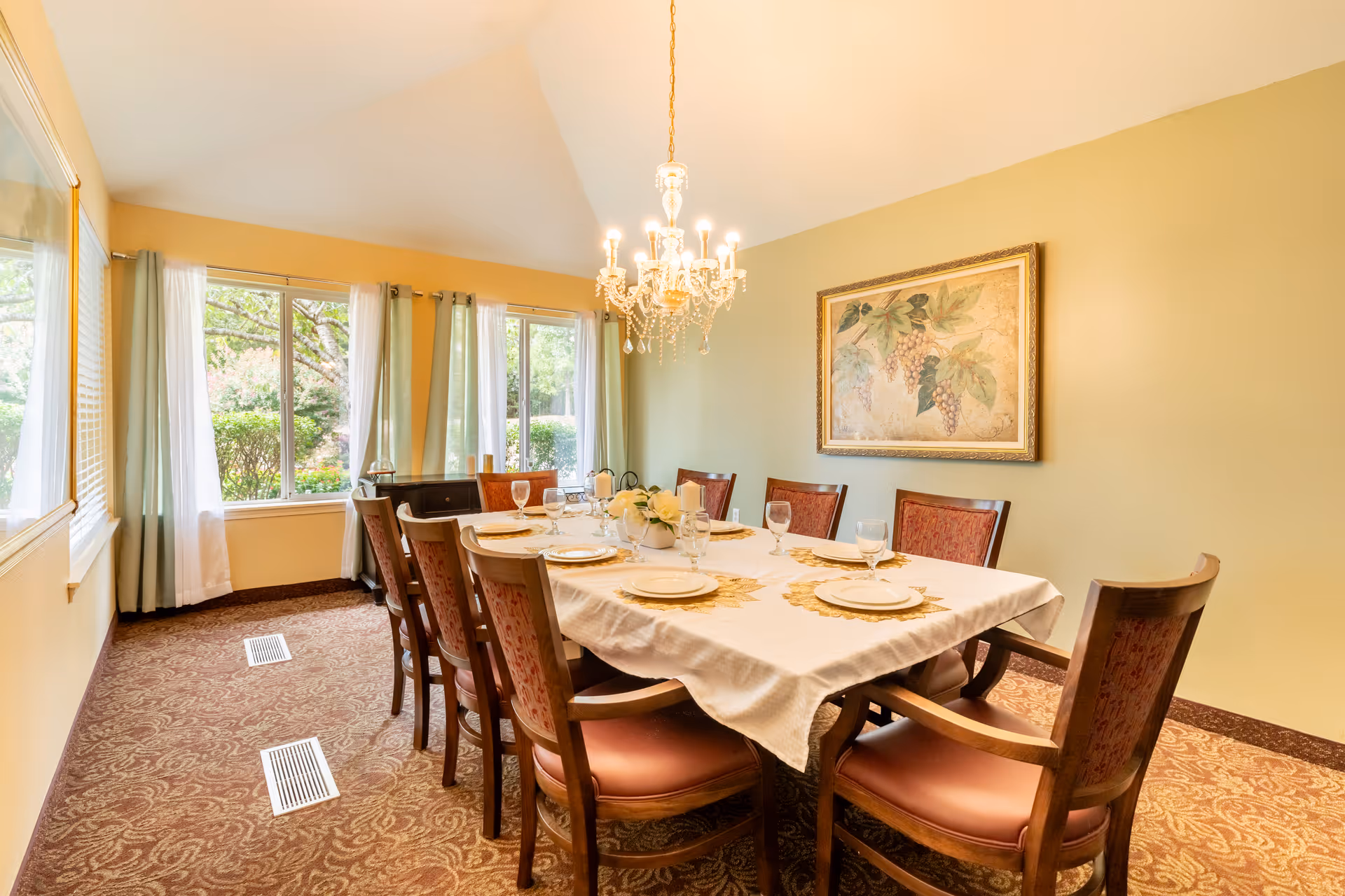 A well-decorated dining room featuring a large table set for a meal, surrounded by elegant chairs. The room has large windows with sheer curtains allowing natural light, and a chandelier hangs from the ceiling. A painting of grapes adorns the wall, adding a touch of sophistication.