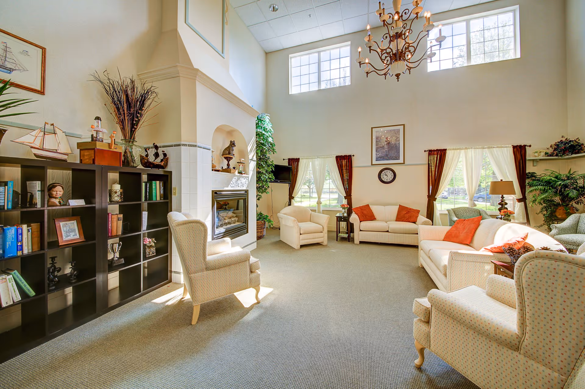 A bright and spacious living room in an assisted living facility with high ceilings and large windows. The room features comfortable upholstered armchairs and sofas with orange accent pillows, a chandelier hanging from the ceiling, a fireplace, a black bookshelf filled with books and decorative items, and several plants. Light filters through the windows with sheer curtains, creating a warm and inviting atmosphere.