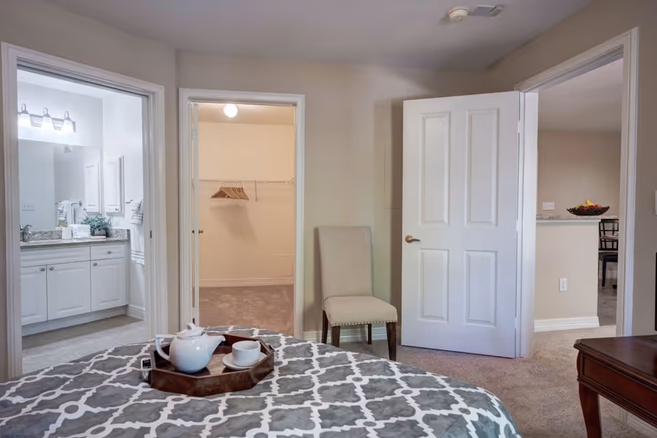 View from a bedroom showing a bed with a patterned gray and white bedspread and a tray with a teapot and cup. The room has an open door leading to a bathroom with a vanity and mirror, another open door leading to a walk-in closet with hangers, and a third open door leading to a hallway with a chair and a glimpse of a dining area.