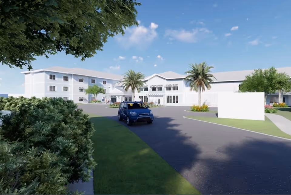 Exterior view of a senior living facility with a driveway, palm trees, bushes, and a blue car driving towards the entrance under a clear blue sky.
