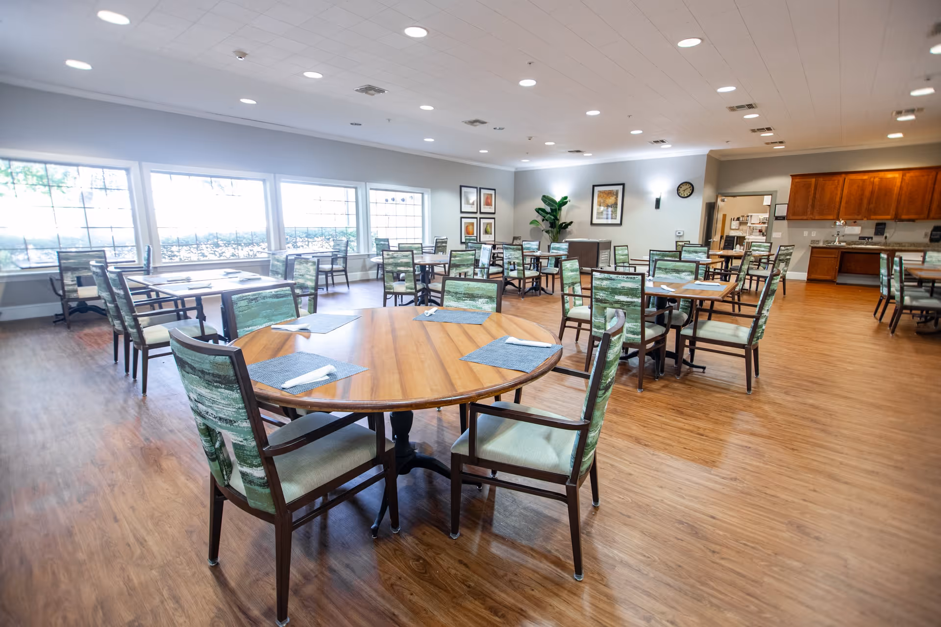 A spacious dining room with multiple wooden tables and chairs arranged neatly. Each table has placemats and napkins set on it. Large windows on one side let in natural light, and the room features wood flooring, light-colored walls, framed artwork, and a clock. A kitchen area with wooden cabinets is visible in the background.