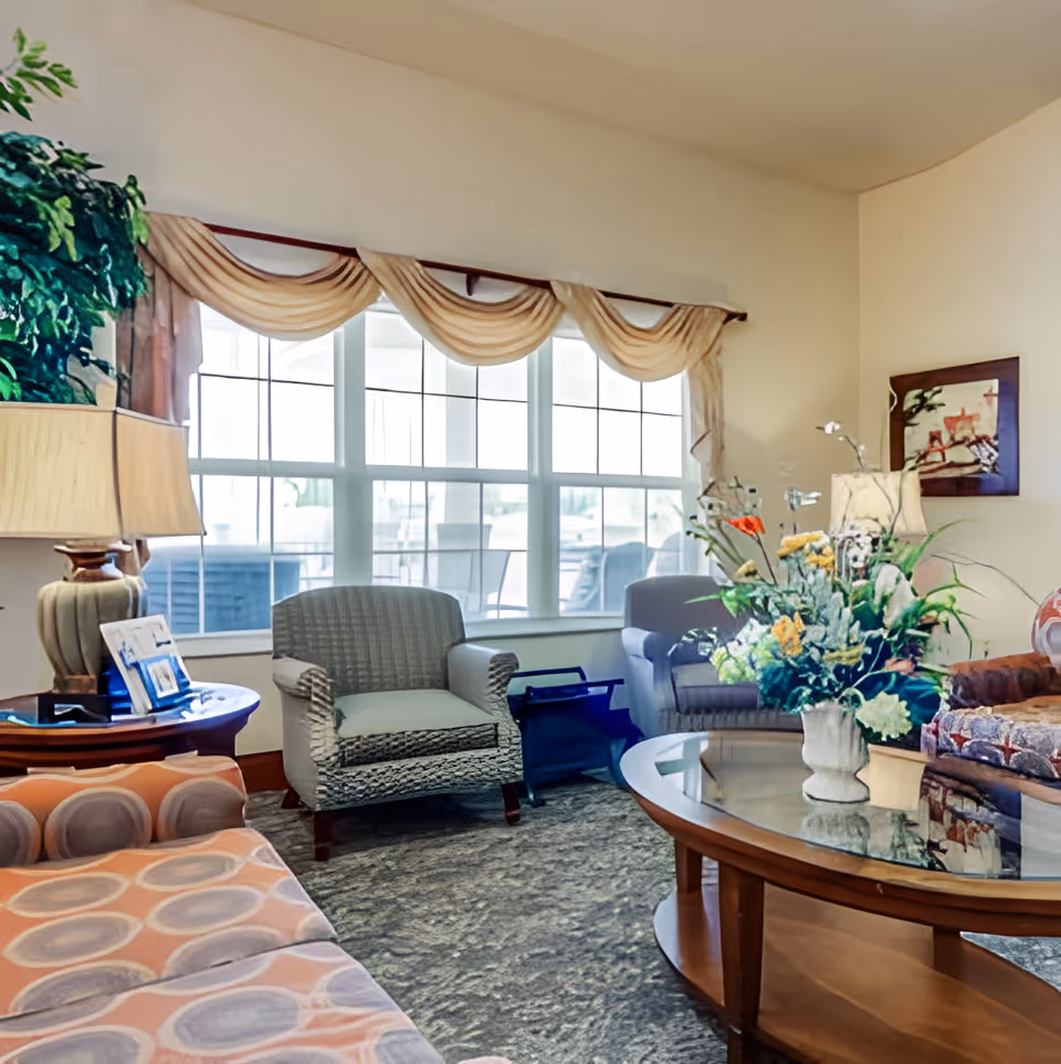 Sunlit communal living room with armchairs and sofas arranged around a glass-topped coffee table topped with a floral arrangement, a lamp, and a large window with drapery.