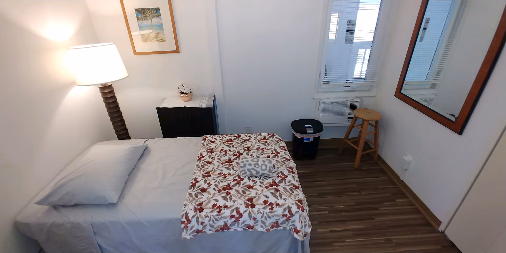 A small, simple bedroom with a single bed covered with a gray sheet and a floral blanket. A pillow is placed at the head of the bed. Next to the bed is a tall floor lamp and a small black nightstand with a decorative flower pot. On the wall above the nightstand is a framed picture. Across from the bed is a window with blinds, an air conditioning unit, a wooden stool, and a black trash bin. A large mirror is mounted on the wall to the right. The floor is wooden.