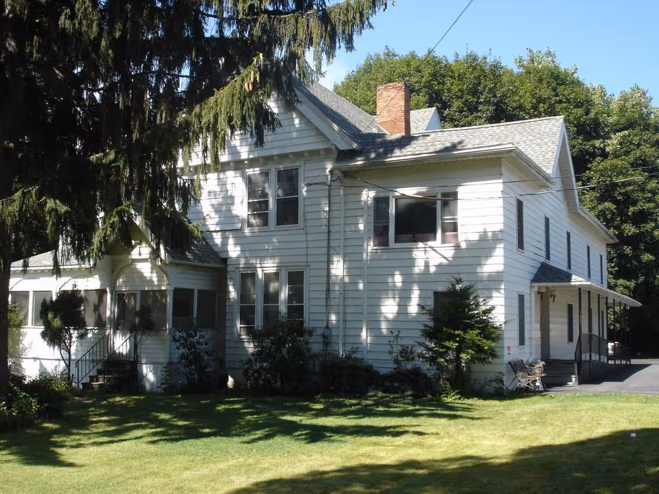 White two-story house with a covered porch, screened entry and a lawn shaded by large trees.