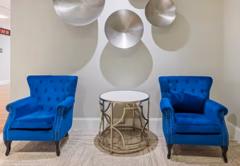 Two blue tufted armchairs flanking a round glass-topped side table beneath metallic circular wall art in a lobby seating area.