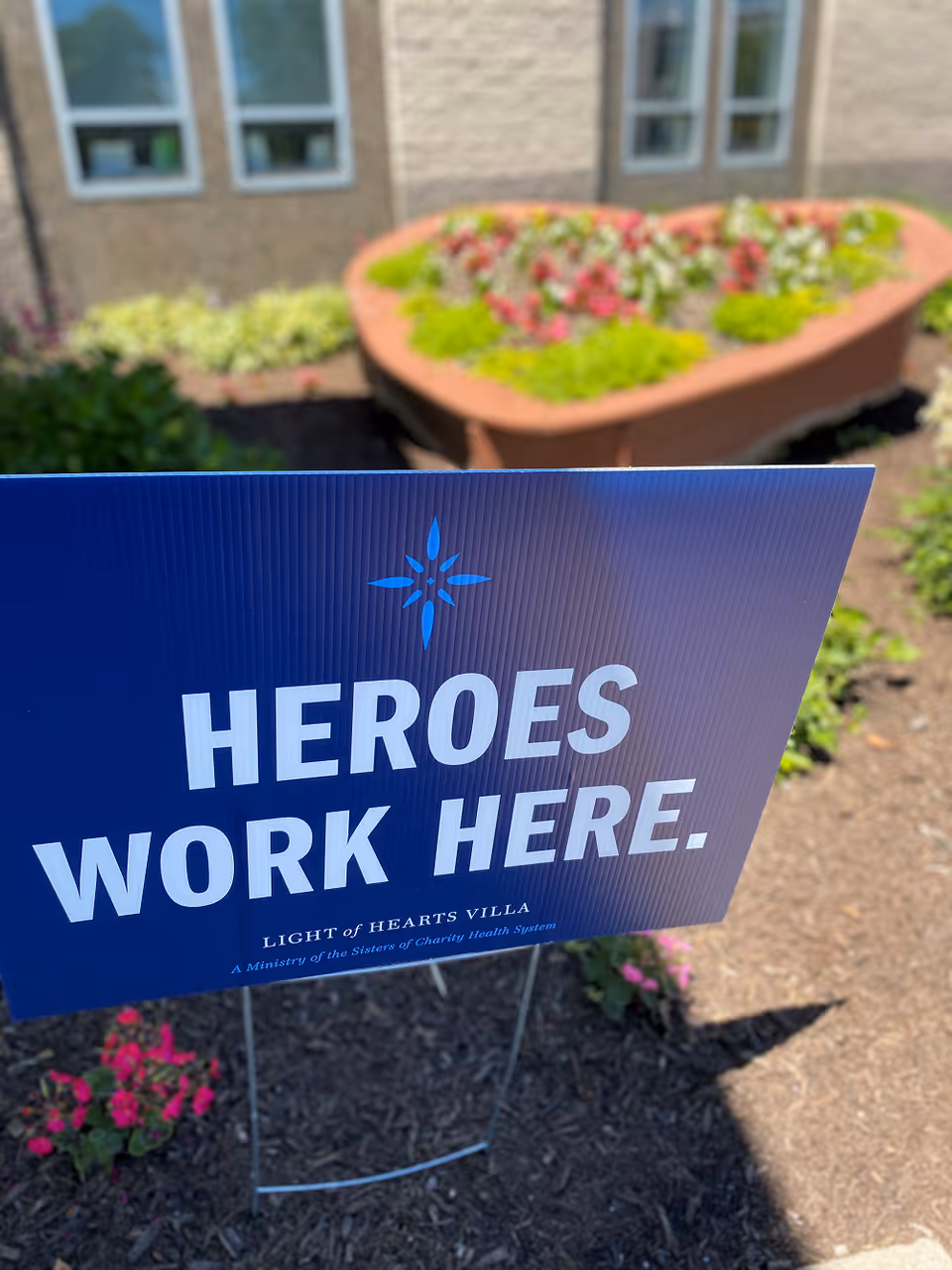 A blue yard sign in a garden bed with flowers and mulch in front of a building. The sign reads 'HEROES WORK HERE.' with 'LIGHT of HEARTS VILLA' and 'A Ministry of the Sisters of Charity Health System' written below.