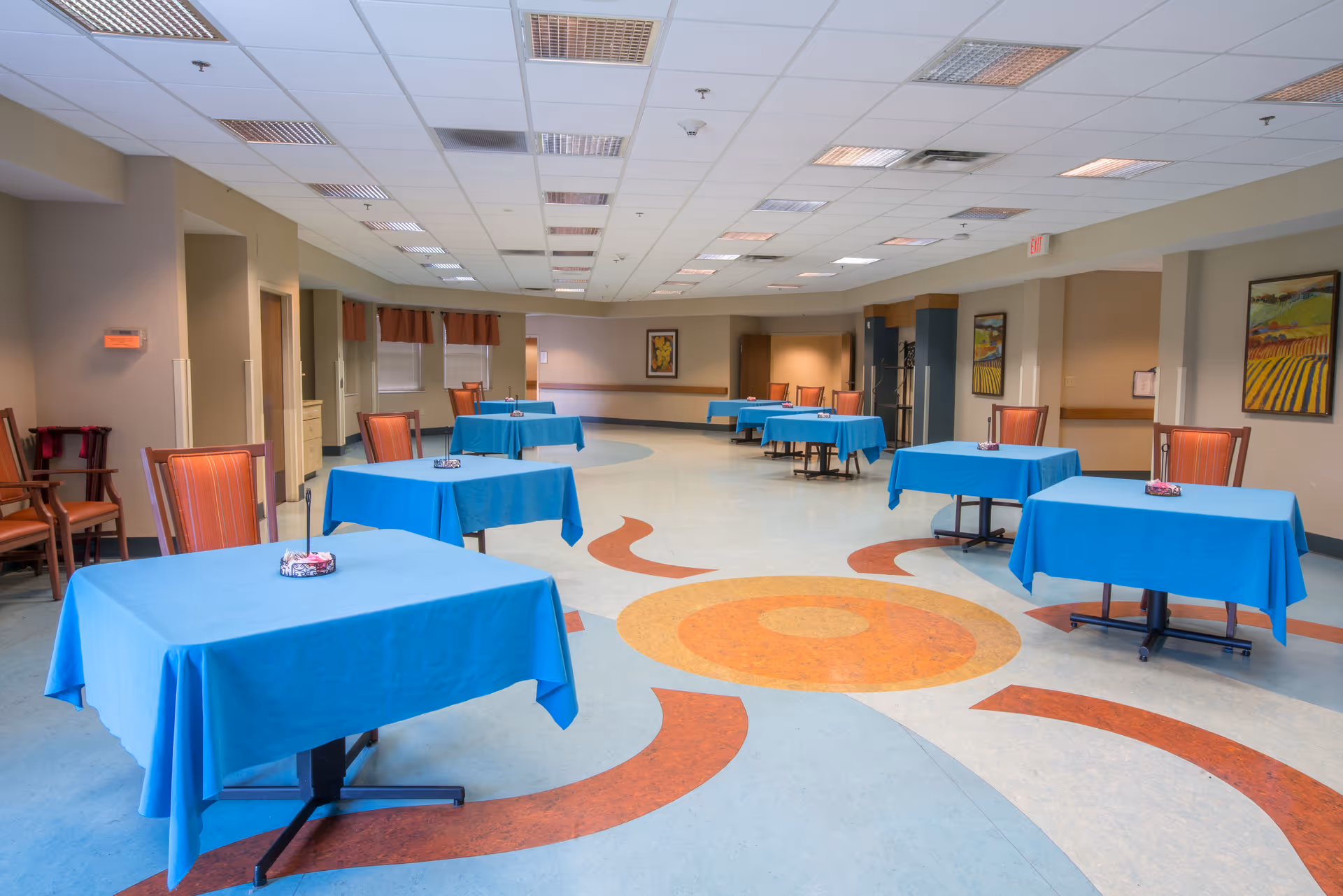A spacious dining area in a senior living facility with several tables covered in blue tablecloths, each accompanied by orange-striped chairs. The floor features a colorful circular pattern, and the walls are decorated with framed artwork. The ceiling has multiple recessed lights, and the room appears clean and well-lit.