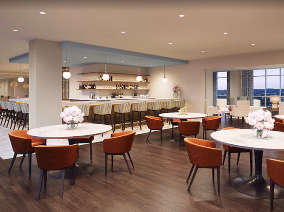 A spacious senior living common area with round tables and orange chairs, a bar with high stools, shelves with bottles and glasses, pendant lights, and large windows with a view outside.