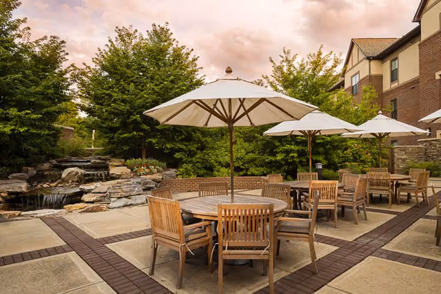 Outdoor patio area with round wooden tables and chairs under large white umbrellas, surrounded by trees and a small waterfall feature, adjacent to a multi-story brick building.