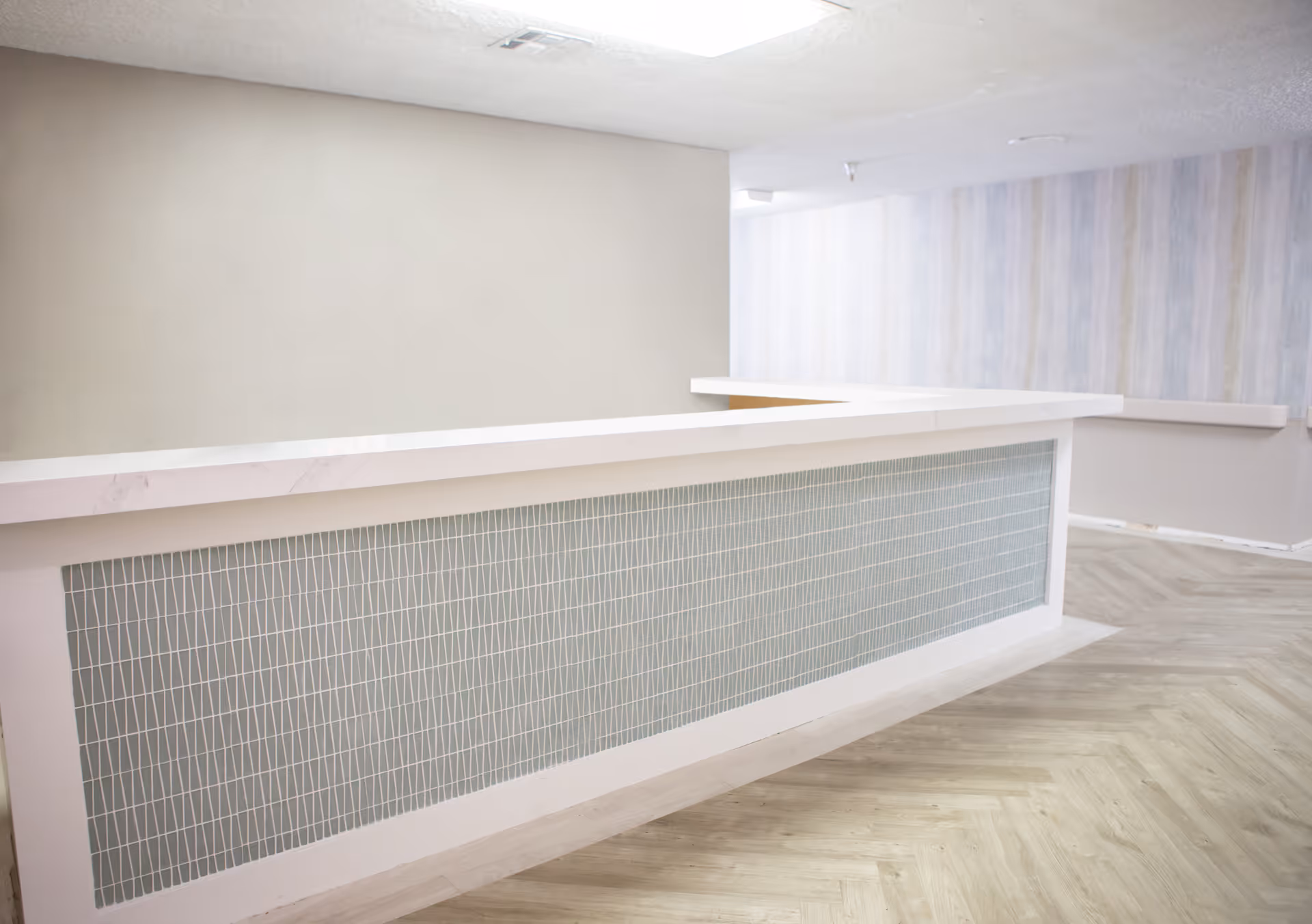 A modern reception desk with a white countertop and a front panel featuring a geometric pattern. The surrounding area has light-colored walls and wood-patterned flooring, creating a clean and welcoming interior space.