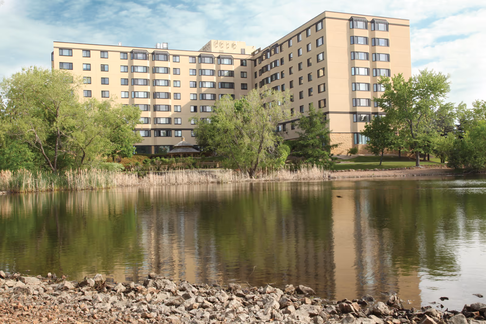 A multi-story beige building with numerous windows is situated behind a pond with a rocky shore. Trees and greenery surround the building and pond, reflecting in the water under a partly cloudy sky.