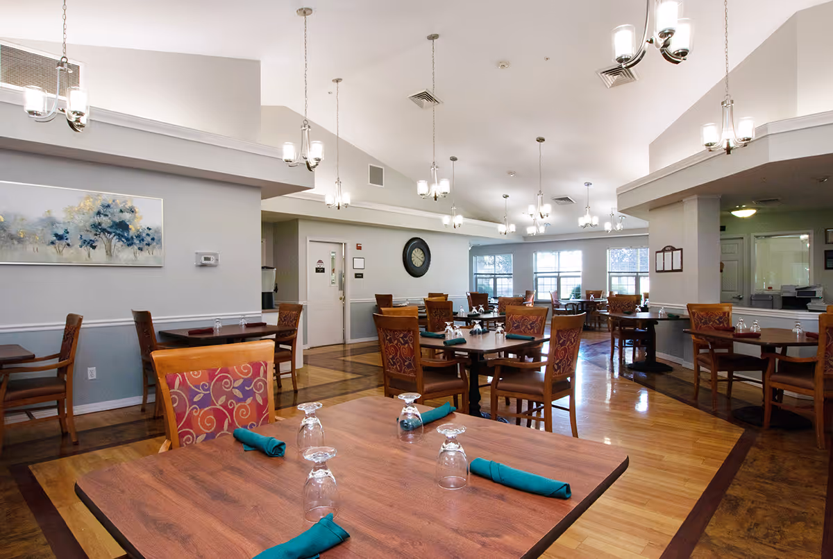A spacious dining room in a senior living facility with multiple wooden tables and chairs. Each table is set with upside-down glasses and green napkins. The room has high ceilings with modern hanging light fixtures, large windows letting in natural light, and a wall clock. The walls are painted in light colors with some artwork displayed.