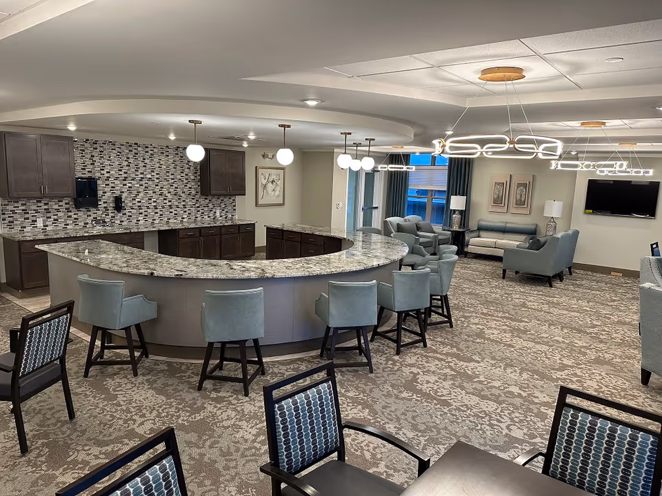 A spacious and modern common area in a senior living facility featuring a large curved granite countertop with several light blue upholstered chairs around it. Behind the counter is a kitchen area with dark wood cabinets and a mosaic tile backsplash. The room also includes multiple seating areas with sofas and armchairs, contemporary light fixtures hanging from the ceiling, and a wall-mounted flat-screen TV.