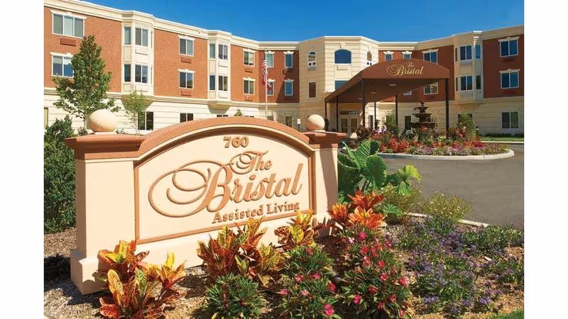 Exterior view of The Bristal Assisted Living at East Northport building with a large sign in the foreground surrounded by colorful flowers and greenery. The building is three stories tall with a brick and beige facade, and an entrance canopy with the facility name. An American flag is visible near the entrance.