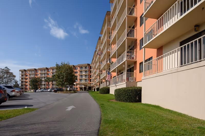 Multi-story apartment building with balconies, a driveway leading to the entrance, parked cars, and American flags under a blue sky.