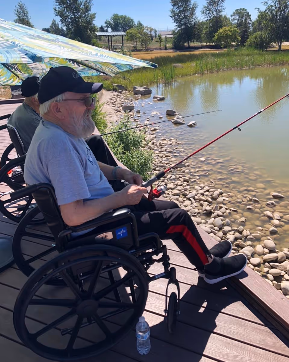 Two elderly men sitting in wheelchairs on a wooden dock by a pond, fishing with rods. One man is wearing a black cap and glasses, and there is a colorful umbrella providing shade. The pond has rocks along the edge and trees in the background under a clear sky.