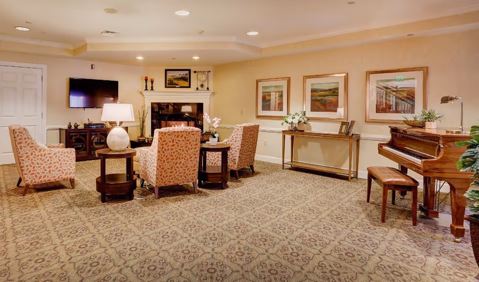 A cozy living room area in a senior living facility featuring four patterned armchairs arranged around two small round tables with a lamp and a plant. There is a fireplace with decorative items on the mantle, a wall-mounted flat-screen TV, framed artwork on the walls, a console table with flowers and picture frames, and a wooden piano with a bench. The room has patterned carpet and soft lighting.