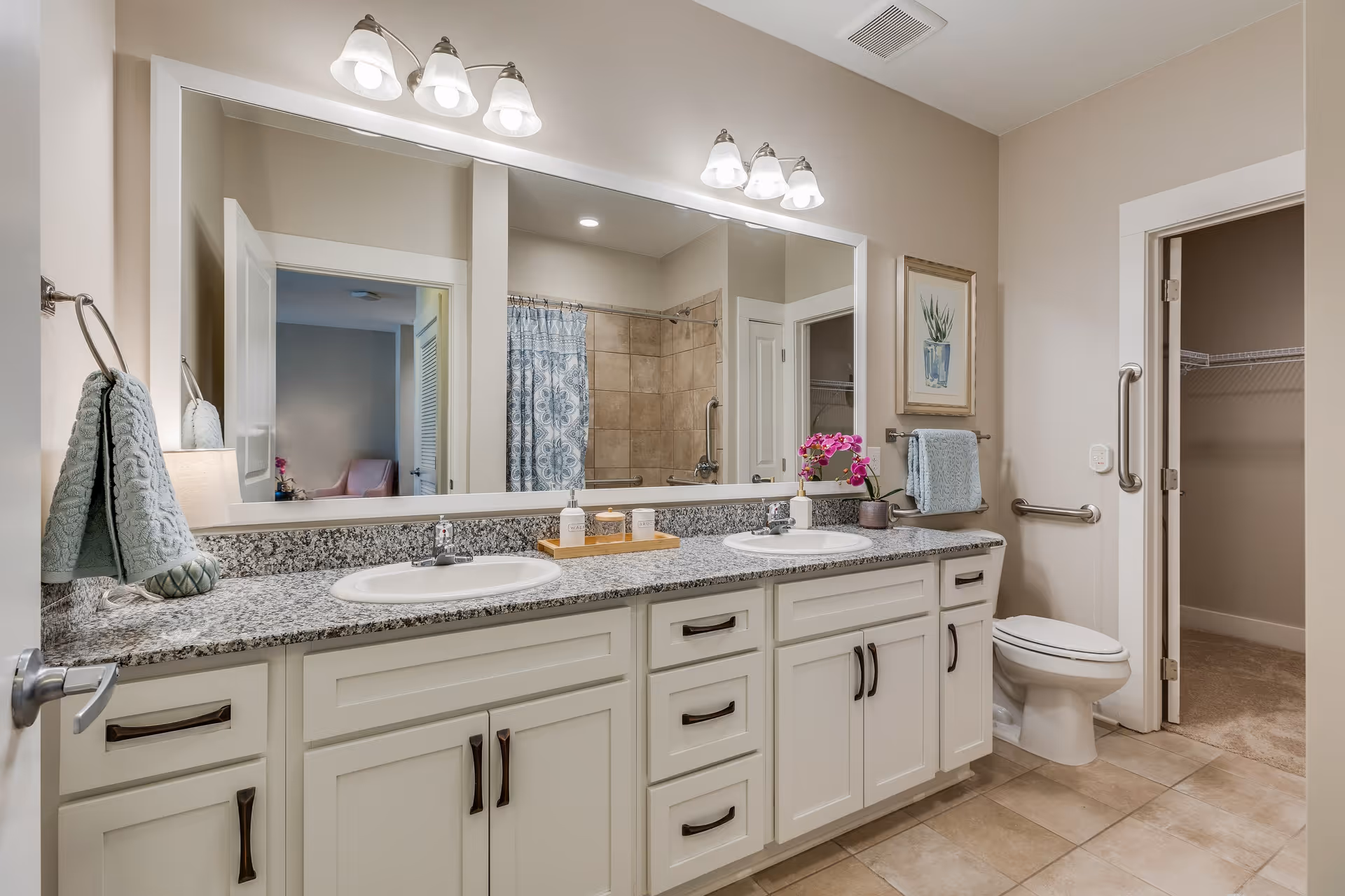 A spacious bathroom featuring a double sink vanity with granite countertop, two mirrors with overhead lighting, a toilet, and a shower with a patterned curtain. The bathroom has beige walls, tiled floor, and a towel rack with a blue towel. There is a framed plant artwork on the wall and an open door leading to a closet.