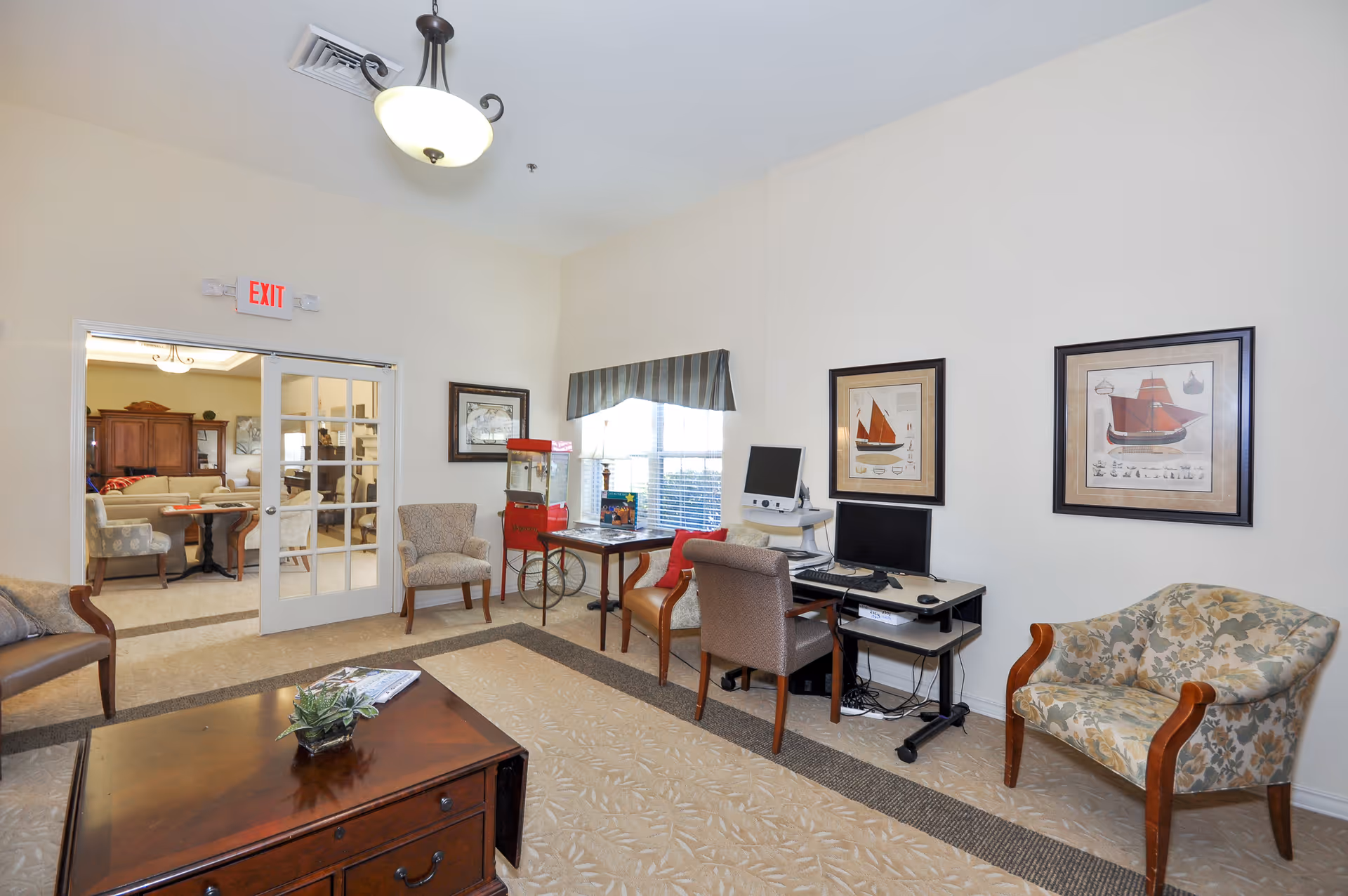 A cozy common area in a senior living facility with several upholstered chairs, a wooden coffee table with a small plant and magazines, a computer workstation, framed nautical-themed artwork on the walls, a window with a striped valance, and a red popcorn machine near the window. Double glass doors lead to another room with more seating and furniture.