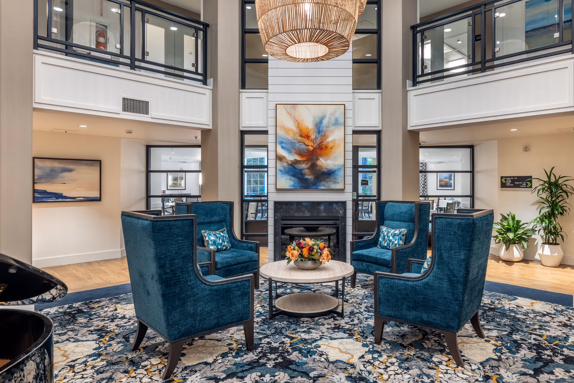A spacious and elegant senior living facility common area with four teal upholstered armchairs arranged around a round coffee table with a floral centerpiece. The room features a large abstract painting above a fireplace, a patterned area rug, potted plants, and a large woven pendant light hanging from the ceiling. There are windows and doors leading to other rooms, and a second-floor balcony with glass railings.