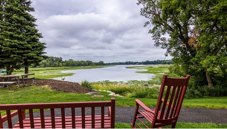 View of a peaceful lake surrounded by greenery under a cloudy sky, with a red wooden bench and rocking chair in the foreground on a grassy area next to a paved path, and a picnic table to the left.