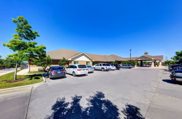 Exterior view of Hunters Pond Rehabilitation and Healthcare Center showing a single-story building with a covered entrance and a parking lot with several parked cars under a clear blue sky.