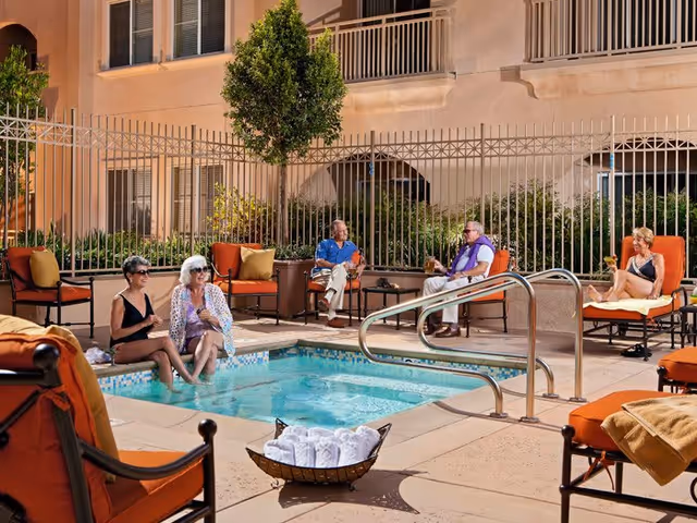 Several older adults relax around a small outdoor hot tub/pool in a gated courtyard with orange lounge chairs and towels.