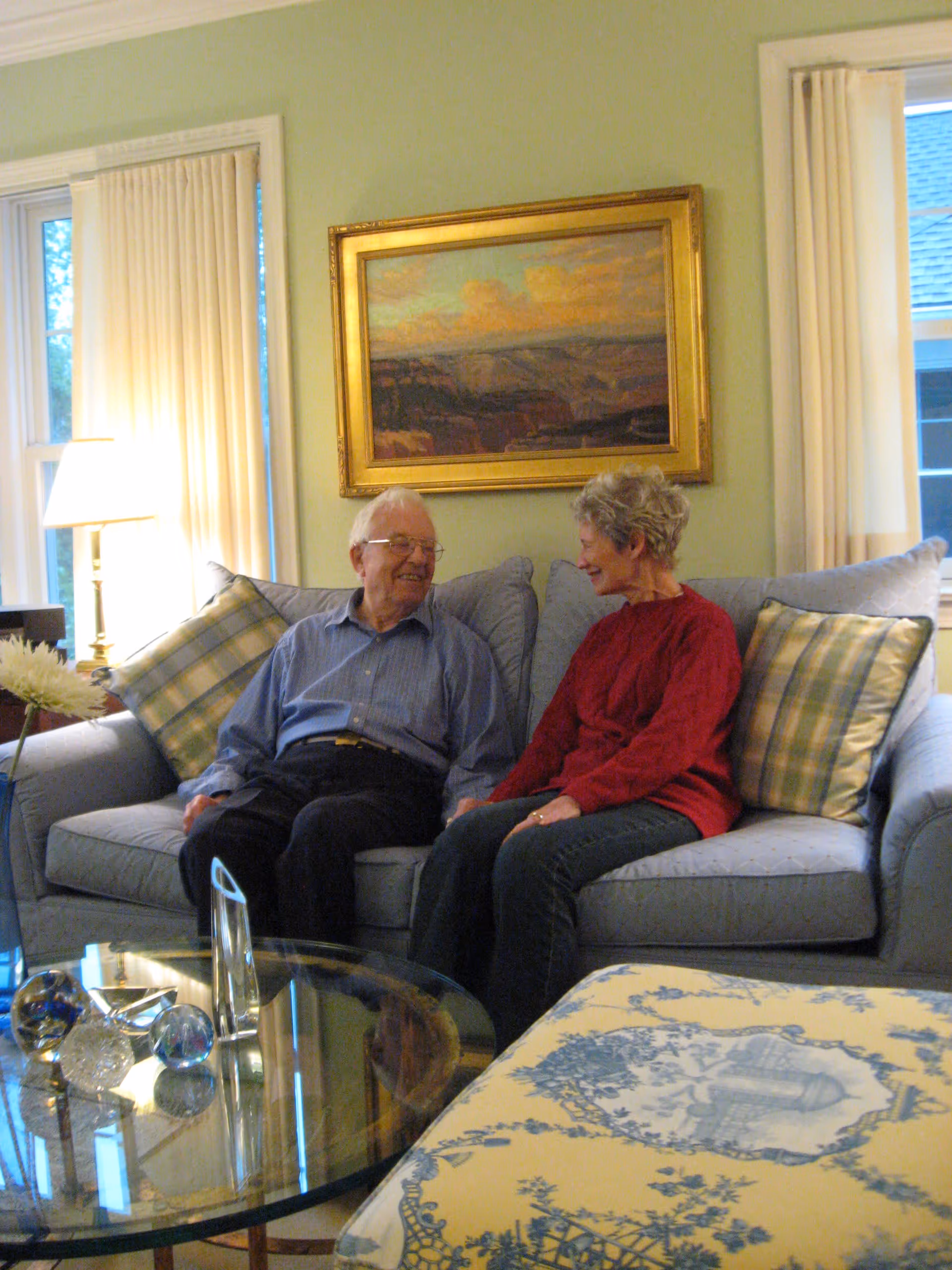 An elderly man and woman sitting on a light blue couch in a cozy living room, smiling and holding hands. The room has light green walls, two windows with cream-colored curtains, a framed landscape painting above the couch, a glass coffee table with decorative glass objects, and a floral patterned ottoman.