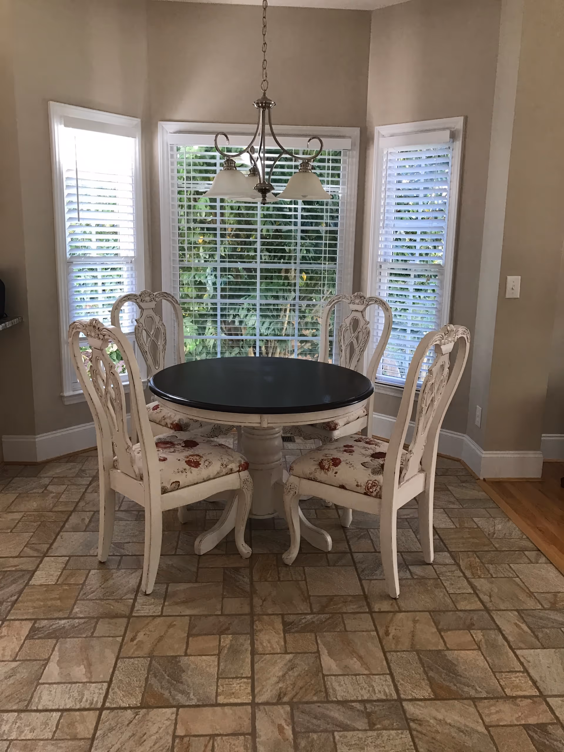A dining area with a round black tabletop on a white pedestal base surrounded by four white wooden chairs with floral upholstered seats. The area has three large windows with white blinds, and a hanging light fixture with three lamps above the table. The floor is tiled with a beige and brown pattern.