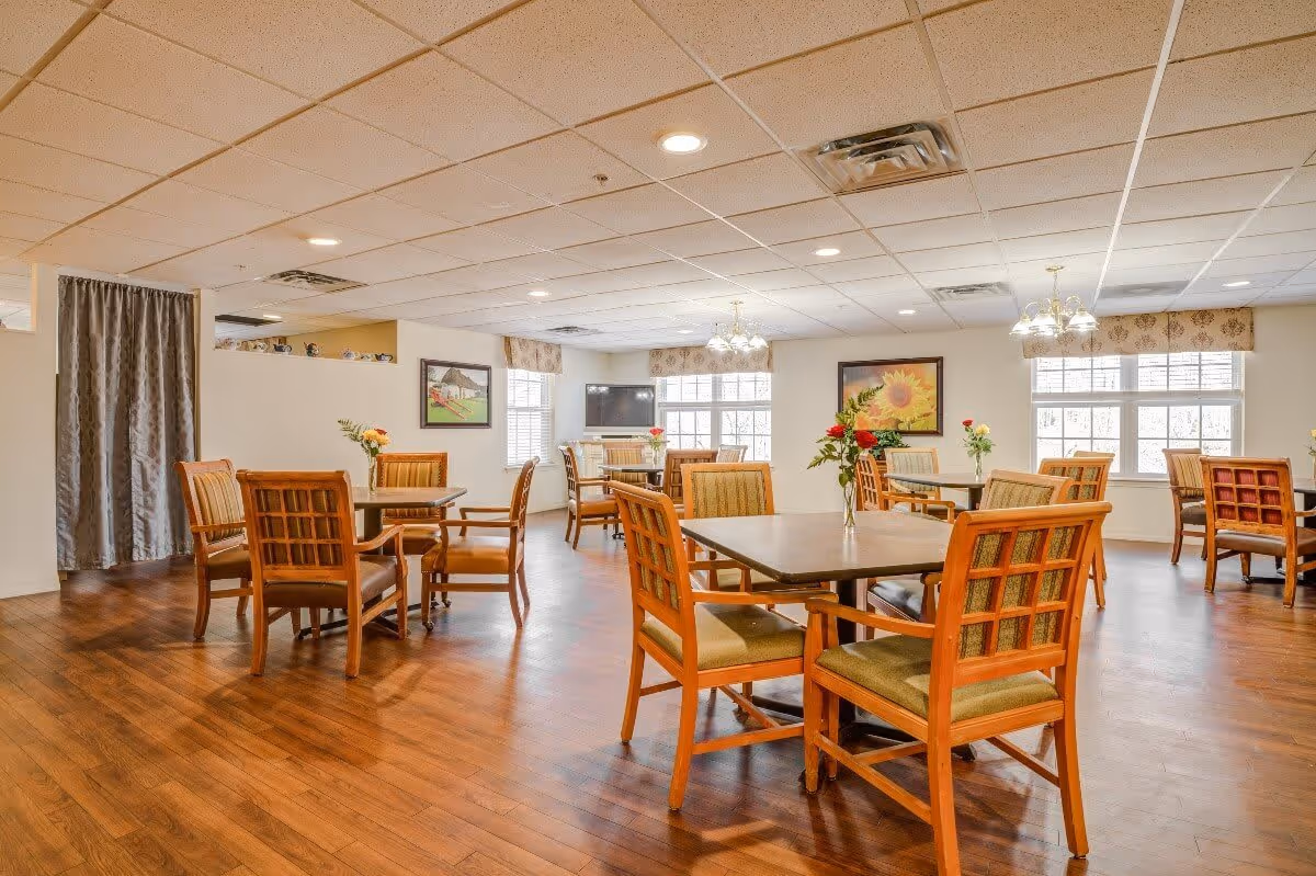 A bright dining room with wooden floors and multiple square tables surrounded by wooden chairs with cushioned seats. Each table has a small vase with flowers. The room has large windows with valances, framed artwork on the walls, and ceiling lights providing illumination.