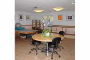 A rehabilitation room with a round table surrounded by four black swivel chairs. The table has a small plant centerpiece. In the background, there is a padded therapy table, exercise ball, and handrails along the walls. Several framed pictures hang on the walls, and a window lets in natural light.