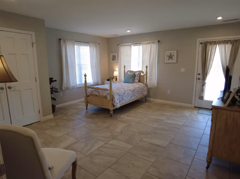 Spacious bedroom with a single wooden bed, tiled floor, windows with curtains, a dresser, and a door to the outside.