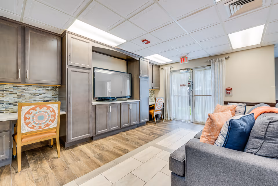Bright communal living area with built-in cabinetry and TV, a sofa with colorful pillows, and glass doors leading outside.
