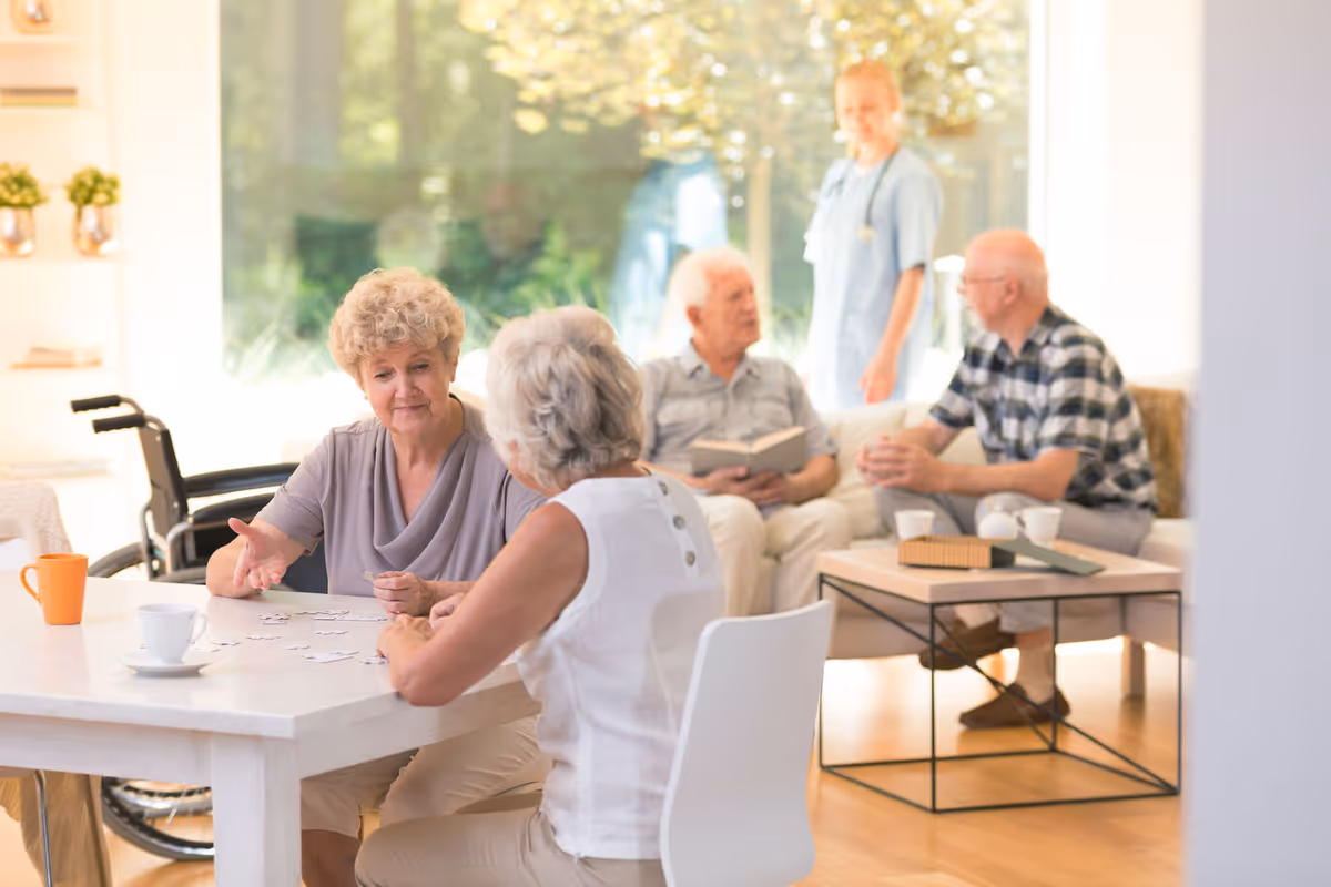 Two elderly women sitting at a table playing a puzzle game with coffee cups nearby, while in the background two elderly men are seated on a couch talking, and a caregiver stands behind them in a bright, spacious room with large windows and natural light.