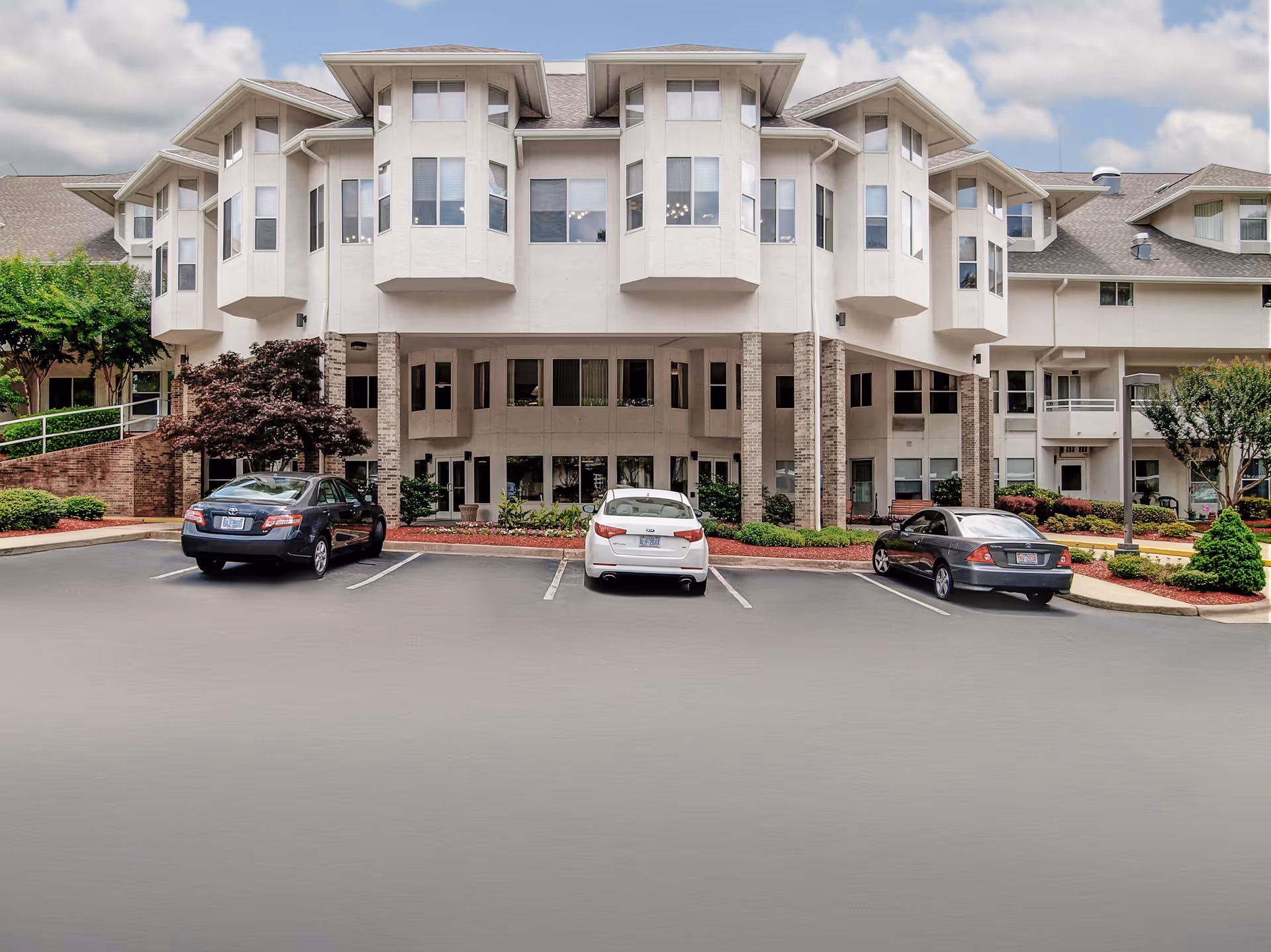 Front exterior view of a multi-story senior living facility building with large windows and architectural details, surrounded by landscaped greenery and a parking lot with three parked cars under a partly cloudy sky.